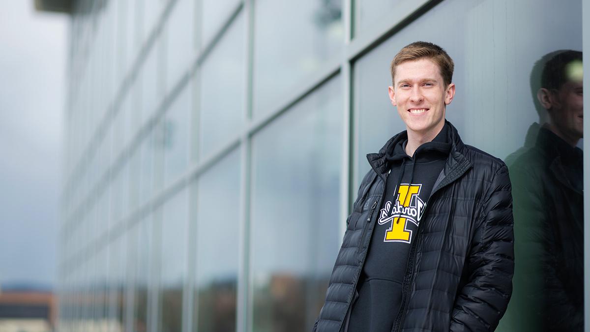 Young man wearing University of Idaho sweatshirt and black jacket leaning against a window at a sport complex.